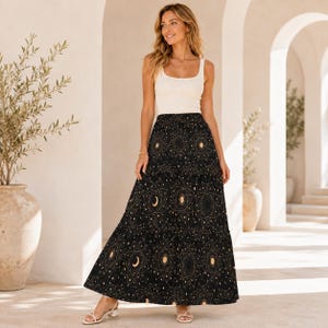May include: A sleeveless white tank top paired with a floor-length black tiered maxi skirt featuring a gold celestial print of moons, stars, and geometric lines. The skirt has a high waist and a loose, flowing silhouette that reaches the ankles.