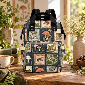 May include: A dark-colored backpack featuring a repeating grid pattern of botanical and wildlife illustrations, including various mushrooms, frogs, and ferns. The bag has black top handles, adjustable shoulder straps, and a front zippered pocket.