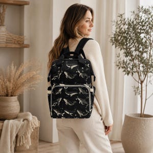 Puede incluir: Mochila negra con estampado de ballenas. La bolsa presenta un diseño repetido de ballenas blancas sobre un fondo negro. Tiene múltiples compartimentos, un asa superior y correas ajustables para los hombros. La bolsa está diseñada para transportar objetos.