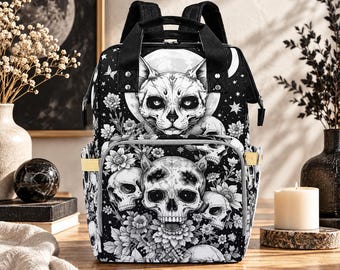 May include: A black and white backpack featuring a detailed graphic print of a cat face centered above a large skull, surrounded by various floral patterns, stars, and crescent moon motifs. The bag includes black top handles and adjustable shoulder straps.