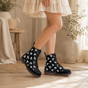 May include: Black combat boots with a white dragon pattern. The boots have a lace-up design and a brown sole. The boots are worn by a person wearing a cream-colored dress.