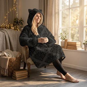 May include: A black hooded blanket with a floral pattern and cat ears. The blanket has a zipper closure and a cream-colored interior. The person is sitting in a chair and holding a white mug.