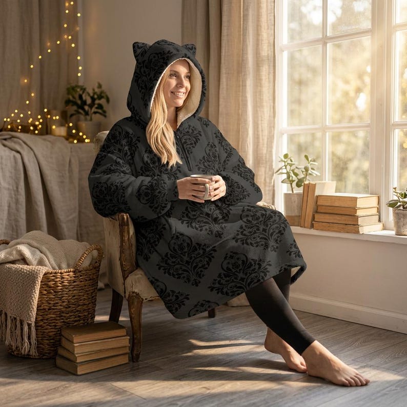May include: A black hooded blanket with a floral pattern and cat ears. The blanket has a zipper closure and a cream-colored interior. The person is sitting in a chair and holding a white mug.