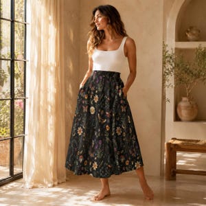 Op de afbeelding: Een wit mouwloos hemdje gecombineerd met een midi-rok met hoge taille op een donkere achtergrond, bedrukt met een kleurrijk patroon van diverse wilde bloemen, groen gebladerte, kleine paddenstoelen en vlinders in een wijd model.