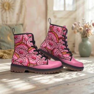 Op de afbeelding: Een paar enkelhoge veterschoenen met een herhalend patroon van roze geglazuurde donuts met kleurrijke hagelslag. De laarzen hebben zwarte veters, zwarte metalen oogjes en een effen roze basis met een dikke, donkerbruine rubberen zool.