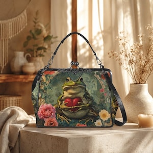 Op de afbeelding: Een rechthoekige handtas met een kikker die een rood hart vasthoudt, omringd door bloemen en bladeren. De tas heeft een zwart handvat en een decoratieve sluiting. Het kleurenpalet omvat groen, roze en een vleugje geel, wat een grillige esthetiek creëert.