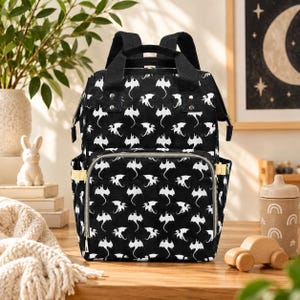 May include: A black backpack featuring a repeating white dragon pattern. The bag includes a large main compartment, a front zippered pocket, two side pockets, and two top carry handles. The design is finished with black straps and silver zippers.