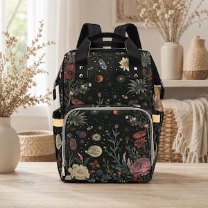 Può includere: Zaino per pannolini con stampa floreale nera con più tasche e una maniglia superiore. La borsa presenta un design celeste e botanico con fiori, pianeti e stelle. La borsa ha spallacci regolabili e una tasca frontale con cerniera.