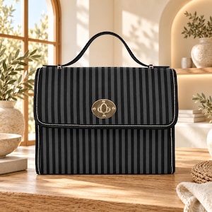 May include: A rectangular handbag featuring a vertical black and dark gray striped pattern. The bag has a single top handle, a fold-over flap, and a central gold-toned metal turn-lock closure. The edges are finished with light-colored stitching.