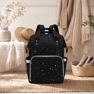May include: Black diaper bag with a colourful speckled pattern. The bag has a large front pocket with a white trim and a zip closure. It features two top handles and adjustable shoulder straps. Designed for carrying baby essentials.