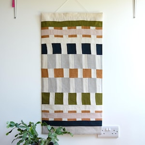 Könnte beinhalten: Ein rechteckiger Wandbehang aus Stoff mit einem geometrischen Muster aus horizontalen und vertikalen Blöcken in Olivgrün, Creme, Hellrosa, Dunkelmarineblau, Hellgrau und Hellbraun. Das Textil ist an einer transparenten Schnur an der Wand befestigt.