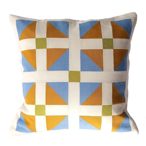 May include: Square decorative pillow with a geometric pattern. The design features squares, triangles, and rectangles in shades of blue, gold, green, and white. The pillow is a square shape.