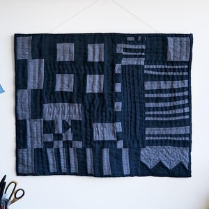 Puede incluir: Un tapiz de pared acolchado negro y gris con un patrón geométrico. El tapiz se muestra en una pared blanca sobre un escritorio con una superficie negra.