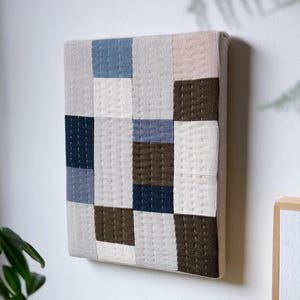 Puede incluir: Colgante de pared cuadrado y acolchado con un diseño de patchwork. La pieza presenta cuadrados de varios colores, incluyendo azul, gris, marrón y blanco. La tela está cosida con costuras visibles. La obra de arte está montada en una pared blanca.