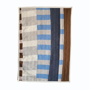 May include: A rectangular quilted textile featuring vertical panels of varying widths. The design includes alternating stripes of brown, cream, light gray, bright blue, and dark navy blue, all held together by visible white running stitch quilting.