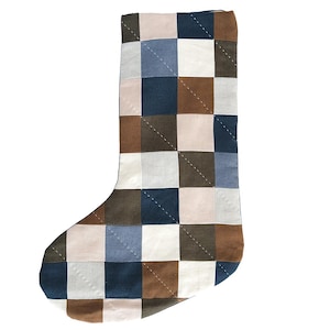 Op de afbeelding: Een gewatteerde kerstsok met een patchworkontwerp van vierkante stoffen panelen in tinten marineblauw, bruin, crème en lichtblauw. De sok is geconstrueerd met zichtbare witte diagonale stiksels die over de vierkanten heen lopen.