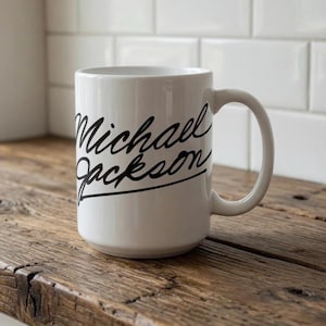 Op de afbeelding: Witte keramische mok met de naam "Michael Jackson" in zwart schrift. De mok heeft een groot oor en staat op een houten oppervlak. De achtergrond bestaat uit witte tegels.