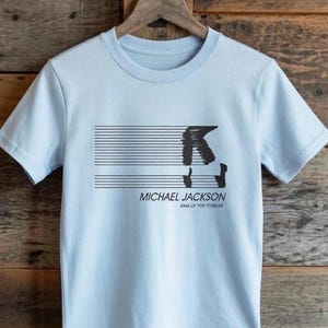 Puede incluir: Camiseta azul claro con un gráfico negro de un bailarín sobre el texto "MICHAEL JACKSON" y "KING OF POP FOREVER". El diseño incluye líneas horizontales. La camiseta está colgada en una percha de madera.