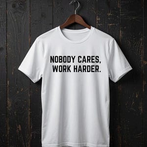Puede incluir: Una camiseta blanca de manga corta con el texto negro "NOBODY CARES, WORK HARDER." impreso en una fuente negrita sin remates en el pecho. La prenda tiene un cuello redondo clásico y se muestra colgada en una percha de madera.
