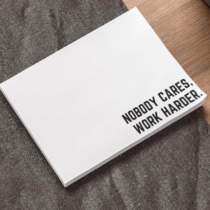 Puede incluir: Un bloc de notas blanco con el texto "NOBODY CARES, WORK HARDER" en letras negras en negrita. El bloc de notas está sobre una superficie de tela gris, con un fondo de madera visible.