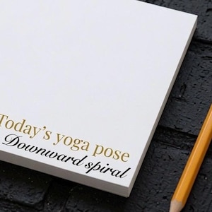 May include: A white notepad with the gold and black text Today's yoga pose Downward spiral printed on the bottom left corner, accompanied by a yellow wooden pencil with a metallic ferrule and a pink eraser tip.