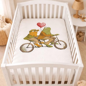 May include: White crib with a fitted sheet featuring a whimsical illustration of two frogs riding a tandem bicycle, holding a red heart-shaped balloon. The room has framed art and a teddy bear.