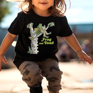 May include: A child wearing a black t-shirt with a green and white graphic of Frog and Toad on a tree branch. The child is wearing patterned pants and black sneakers while riding a skateboard.