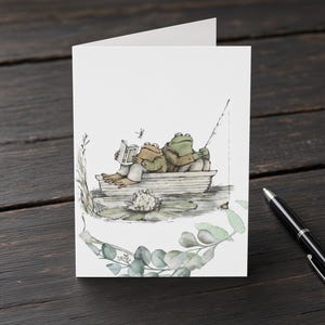 Puede incluir: Una tarjeta de felicitación blanca con una ilustración en acuarela de dos ranas verdes sentadas en un pequeño bote de madera sobre el agua. Una rana lee un libro mientras la otra sostiene una caña de pescar. Un nenúfar blanco y follaje decoran la parte inferior.