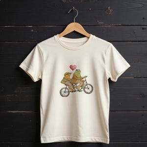 Op de afbeelding: Crèmekleurig T-shirt met een grillige illustratie van twee kikkers op een tandemfiets. De kikkers dragen jassen en hoeden, met een rood hart erboven. Het ontwerp is gedrukt op een klassiek T-shirt met ronde hals.