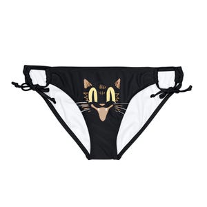 May include: Black bikini bottoms with a cat face graphic. The cat has yellow eyes, a tan nose, and whiskers. The bikini bottoms have white lining and black ties on each side.
