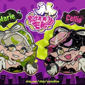 May include: Two enamel pins featuring cartoon characters, Marie and Callie, from the video game Splatoon. Marie is green and white, Callie is black and pink. The pins have a purple background with green splatters and city silhouettes.