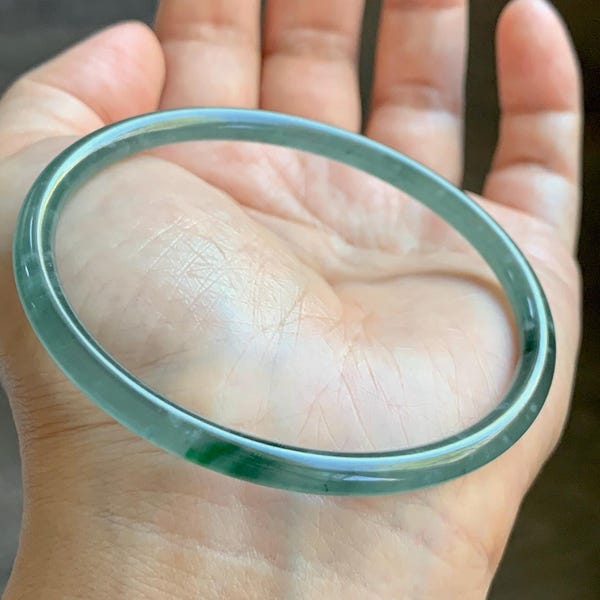 May include: A smooth, translucent, circular bangle bracelet featuring a light green hue with subtle darker green inclusions throughout the polished stone surface, creating a continuous, solid, and rounded band design.