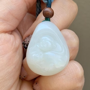 Puede incluir: Colgante de jade blanco tallado con una figura de Buda sonriente. El colgante en forma de lágrima cuelga de un cordón azul con una cuenta de madera. El jade tiene una superficie lisa y pulida.