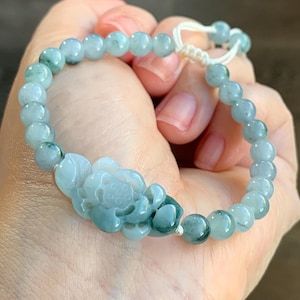 May include: A jade bracelet featuring a carved flower charm. The bracelet is made from light green jade beads and has a white string fastening.
