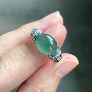 May include: A silver ring featuring a large, oval-shaped, translucent green gemstone. The ring is accented with small, clear gemstones on the band. The ring is held in a hand, showcasing the jewelry's design and color.