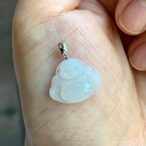 May include: A small, light-colored pendant in the shape of a smiling Buddha. The pendant is made of a translucent material and has a silver-colored bail. The Buddha pendant is held in a hand.