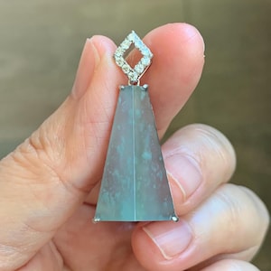 May include: A trapezoidal pendant featuring a translucent, mottled blue-green stone set in a silver-toned frame. The top of the pendant is adorned with a diamond-shaped bail encrusted with small, clear, sparkling stones that catch the light.