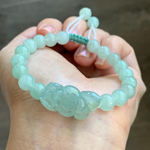 May include: A light green jade bracelet with round beads and a carved lotus flower centerpiece. The bracelet is held in a hand, showcasing its adjustable design with a green and white cord.