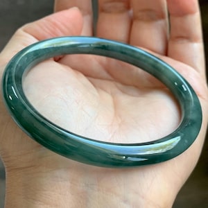 May include: A smooth, circular bangle bracelet crafted from translucent dark green stone. The piece features a polished, glossy finish with subtle variations in color intensity throughout the material, creating a deep, rich appearance in this solid, continuous band.
