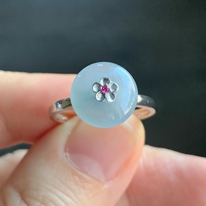 May include: A silver ring with a light blue oval-shaped stone set in the center. The stone has a silver flower design with a small pink stone in the center.