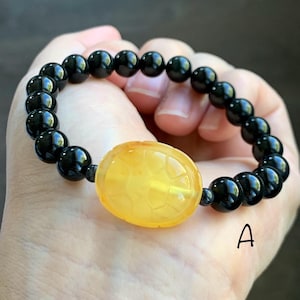 May include: Black beaded bracelet with a large, oval, yellow-orange center stone. The center stone has a carved design. The bracelet is held in a hand. The letter "A" is visible in the image.