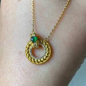 May include: A gold necklace with a circular pendant featuring a green gemstone and small clear stones. The pendant is surrounded by a braided design.