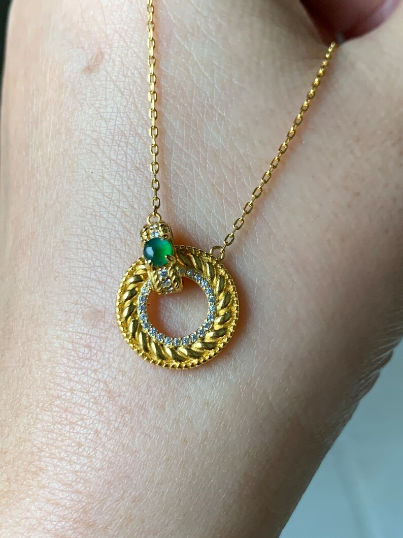 May include: A gold necklace with a circular pendant featuring a green gemstone and small clear stones. The pendant is surrounded by a braided design.