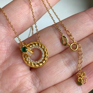 May include: A gold chain necklace with a circular pendant featuring a braided design and a small green gemstone. The pendant is accented with clear rhinestones.