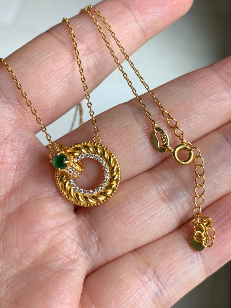 May include: A gold chain necklace with a circular pendant featuring a braided design and a small green gemstone. The pendant is accented with clear rhinestones.