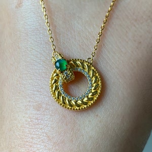 May include: A gold chain necklace with a circular pendant featuring a green gemstone and a braided design accented with small clear stones.
