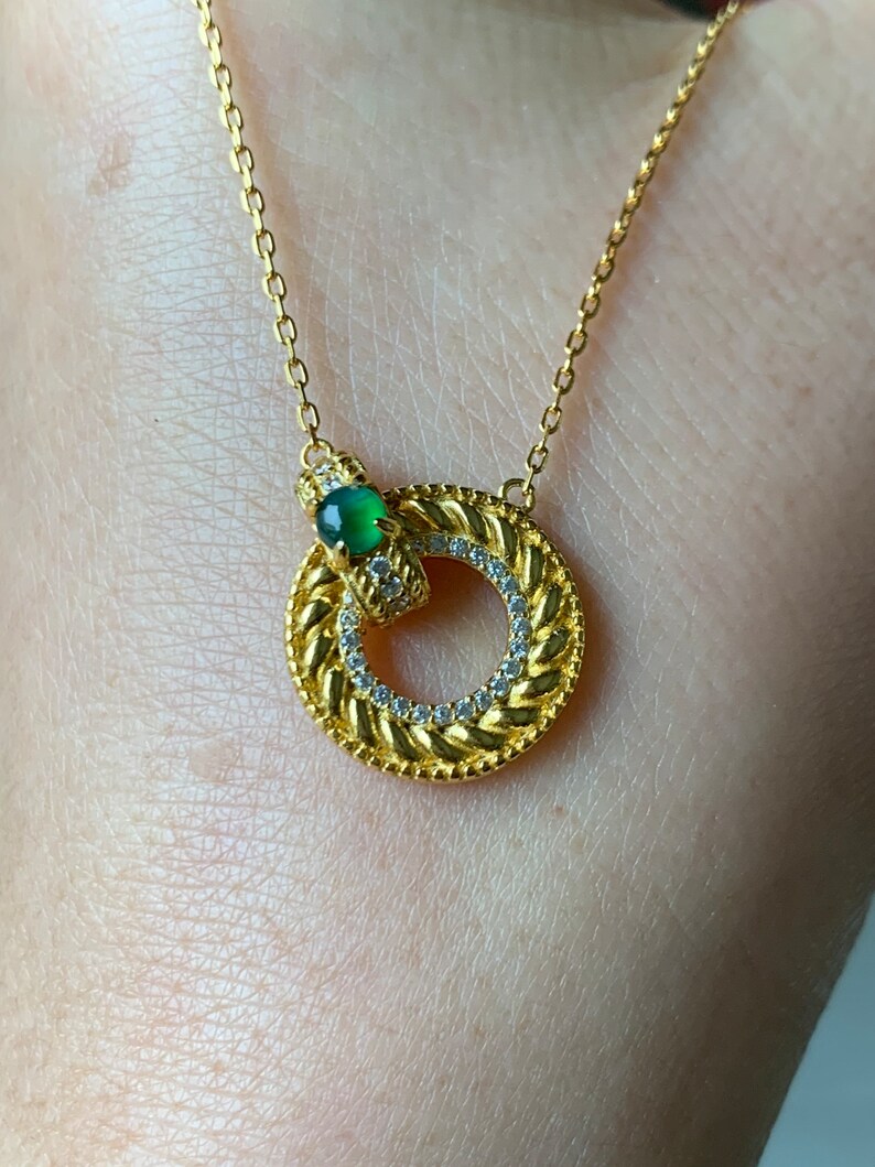 May include: A gold chain necklace with a circular pendant featuring a green gemstone and a braided design accented with small clear stones.