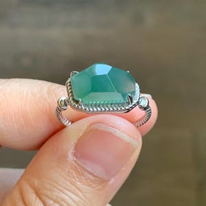 Könnte beinhalten: Ein Silberring mit einem großen, facettierten, teal-farbenen Edelstein. Der Edelstein ist in einer dekorativen, strukturierten Lünette gefasst. Das Band des Rings hat ein gedrehtes Seildesign. Der Ring wird von den Fingern einer Person gehalten.