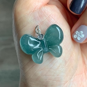 Puede incluir: Un colgante de mariposa de color verde azulado con una fianza plateada. El colgante está hecho de un material translúcido, posiblemente jade o vidrio, y tiene un diseño tallado detallado. Las alas de la mariposa son redondeadas y simétricas.