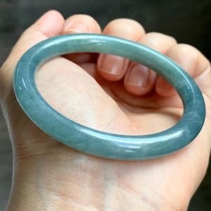 Könnte beinhalten: Ein transluzentes, teal-farbenes Jade-Armreif-Armband. Die glatte, polierte Oberfläche reflektiert das Licht und zeigt die natürlichen Variationen des Steins. Das Armband ist ein vollständiger Kreis, der am Handgelenk getragen werden soll. Der Jade scheint ein durchgehendes Stück zu sein.